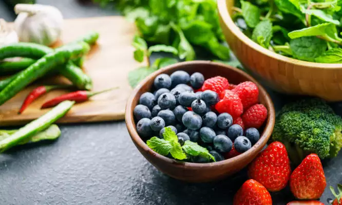 Ένα μπολ με φρέσκα μούρα που περιβάλλεται από πράσινα πράσινα, μπρόκολο, φράουλες και πιπεριές σε μια μαύρη επιφάνεια πάγκου με το προϊόν Αντιοξειδωτικά.