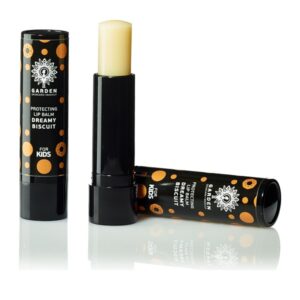 Δύο σωληνάρια Garden Protecting Lip Balm Dreamy Biscuit Kids 5,2 g, το ένα ανοιχτό με βάλσαμο ανοιχτό και το άλλο κλειστό, με μαύρη και πορτοκαλί συσκευασία. Αυτό το βάλσαμο για τα χείλη είναι τέλειο για παιδιά, διατηρώντας τα χείλη τους λεία κατά τις υπαίθριες περιπέτειες.