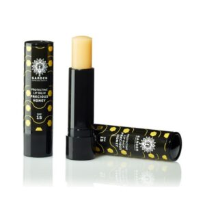 Δύο μαύρα σωληνάρια Garden Protecting Lip Balm Precious Honey SPF15 5,2 g, το ένα ανοιχτό για να αποκαλύψει το βάλσαμο, με σχέδιο κηρήθρας.