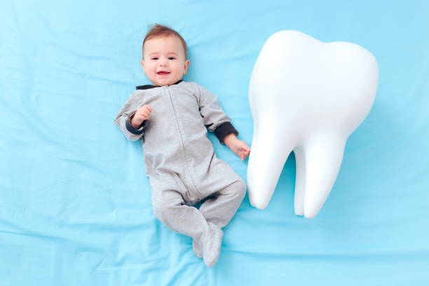 A baby in a gray onesie ξαπλώνει σε μπλε φόντο δίπλα στη Στοματική Υγιεινή Παιδιών, χαμογελώντας και κοιτάζοντας ψηλά.