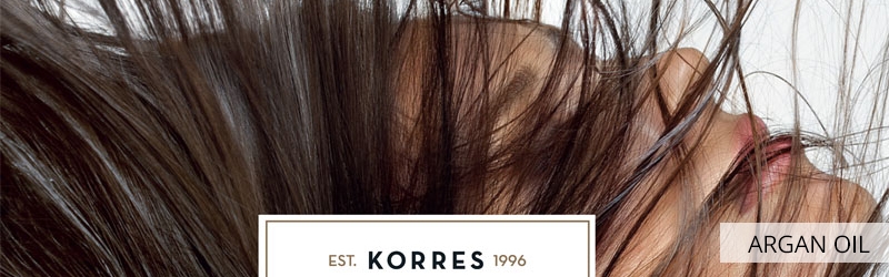 Κοντινό πλάνο ενός ατόμου με μακριά, σκούρα μαλλιά που καλύπτουν το πρόσωπό του. Το κείμενο γράφει "ARGAN OIL" και "EST. KORRES 1996".